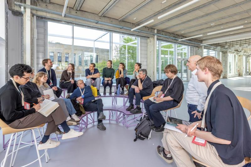 A group of students and professors sit in a circle in a room with big glass windows