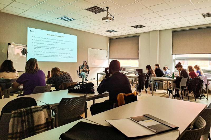 Group of university students in a classroom listen to a presentation by a female lecturer