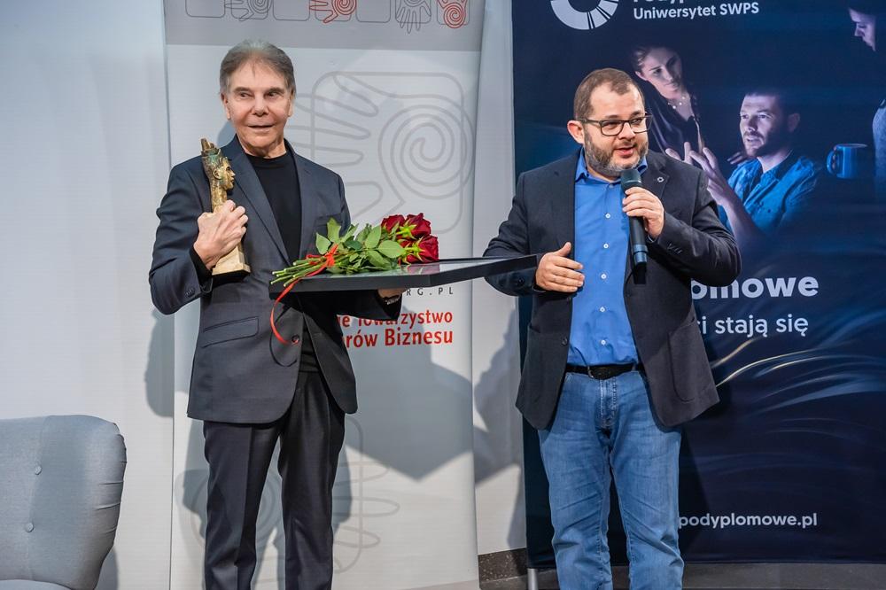 Professor Cialdini holding a statue and flowers, next to him stands Professor Tomasz Grzyb with a microphone