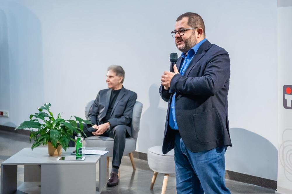 Photo of the panel, Professor Tomasz Grzyb stands on the foreground with a microphone, Professor Cialdini sits in the background