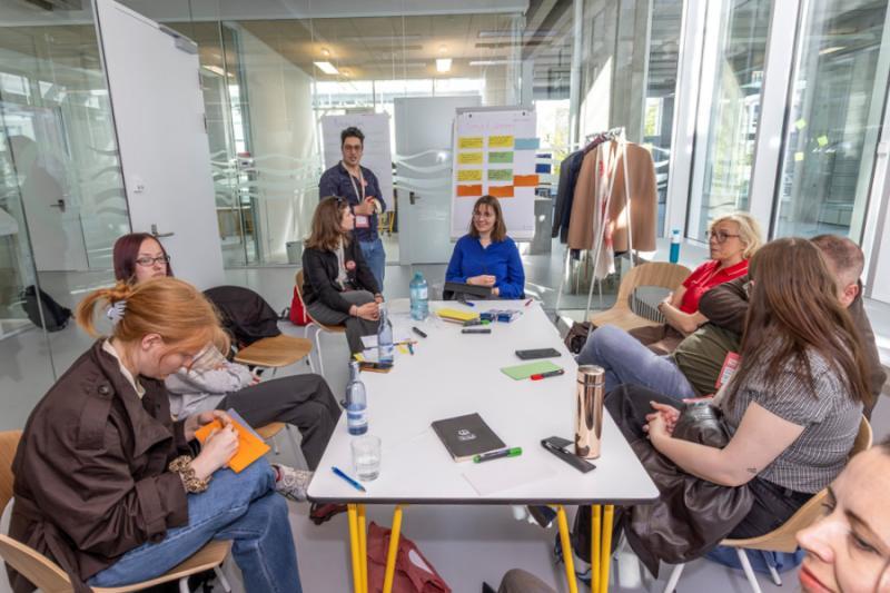 Students gathered around a table listening looking at a flipchart and a presenter