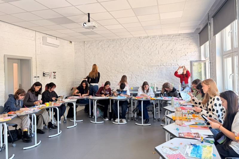 Students sit at desks arranged in a circle and work on their artworks in a classroom