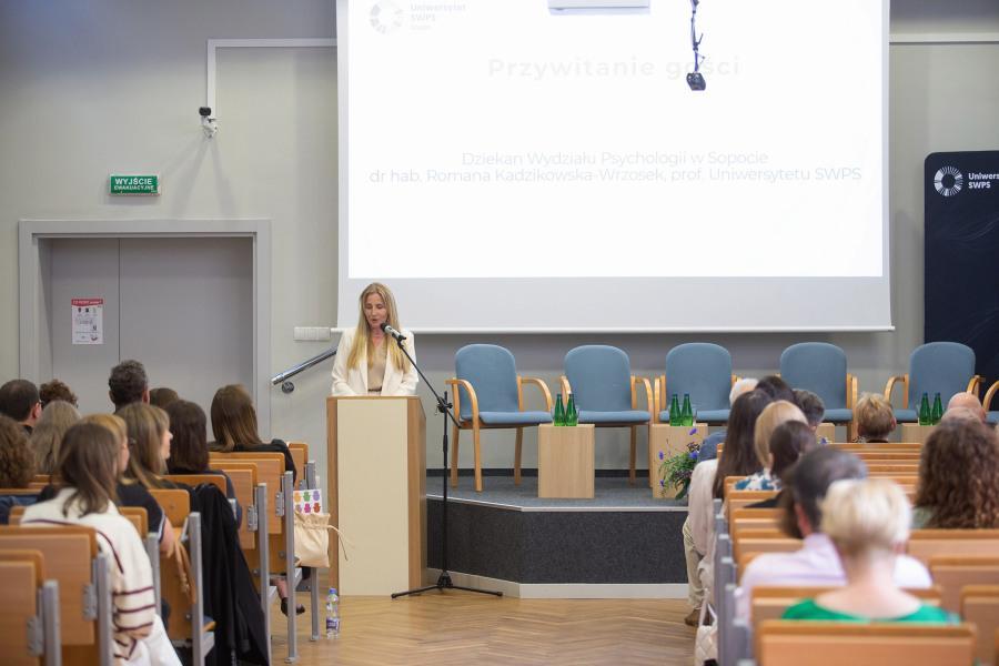 Dean of the Faculty of Psychology in Sopot, SWPS University, Wydziału Psychologii w Sopocie, prof. Romana Kadzikowska-Wrzosek, is giving a speech at a podium in the background. Audience is sitting on chairs and listening in the foreground.