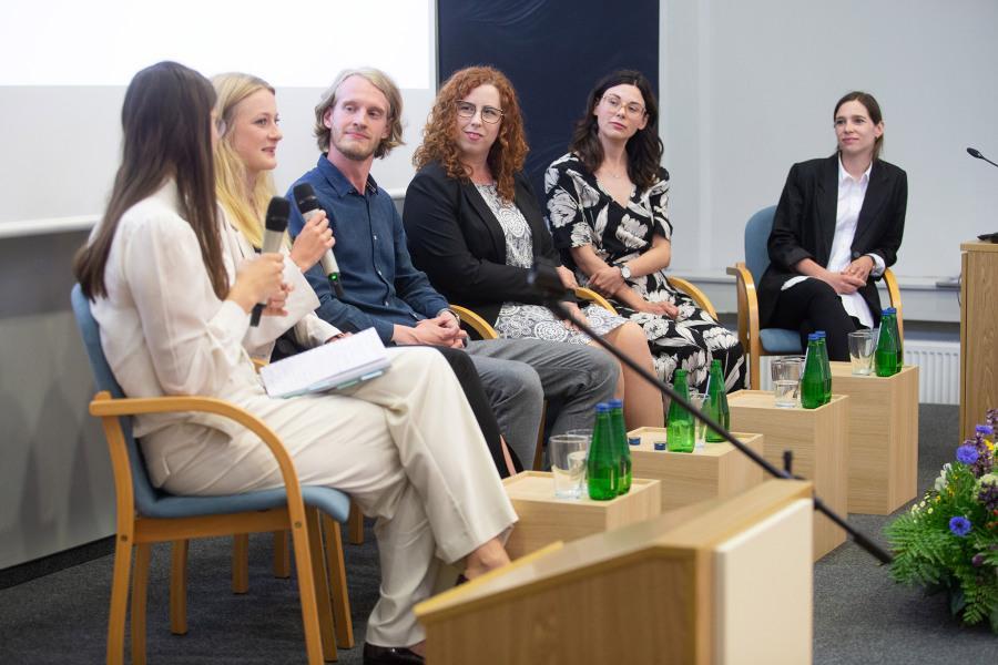 Five graduates from the Faculty, accompanied by a discussion moderator, seated on chairs. Two individuals hold microphones. Tables with water bottles are placed in front of the panelists.