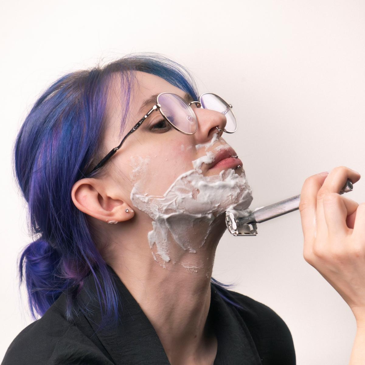 A girl with blue hair is shaving her chin with a metal razor.
