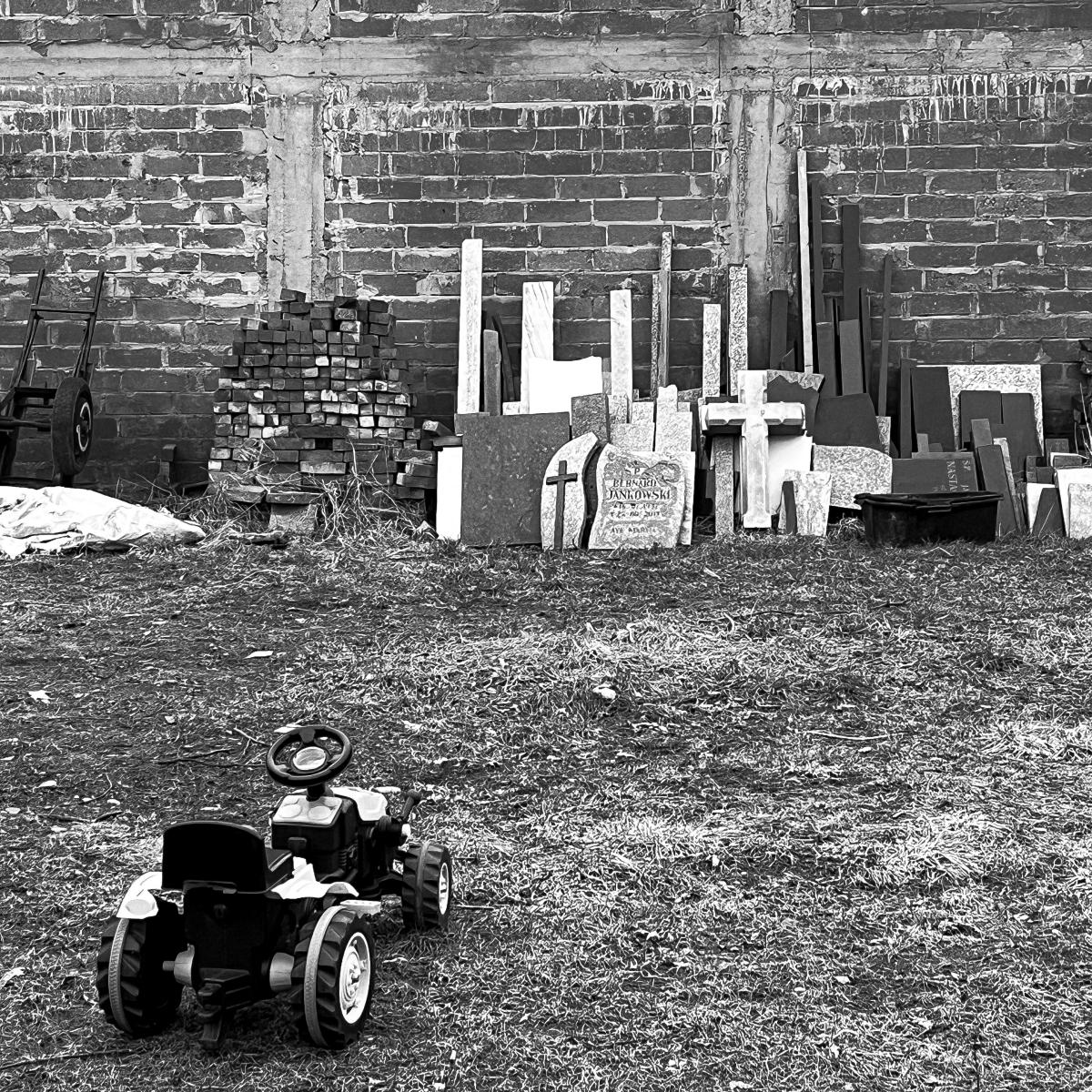 A black-and-white photograph; in the foreground, a children’s ride-on tractor; in the background, crosses and headstones leaning against a wall.