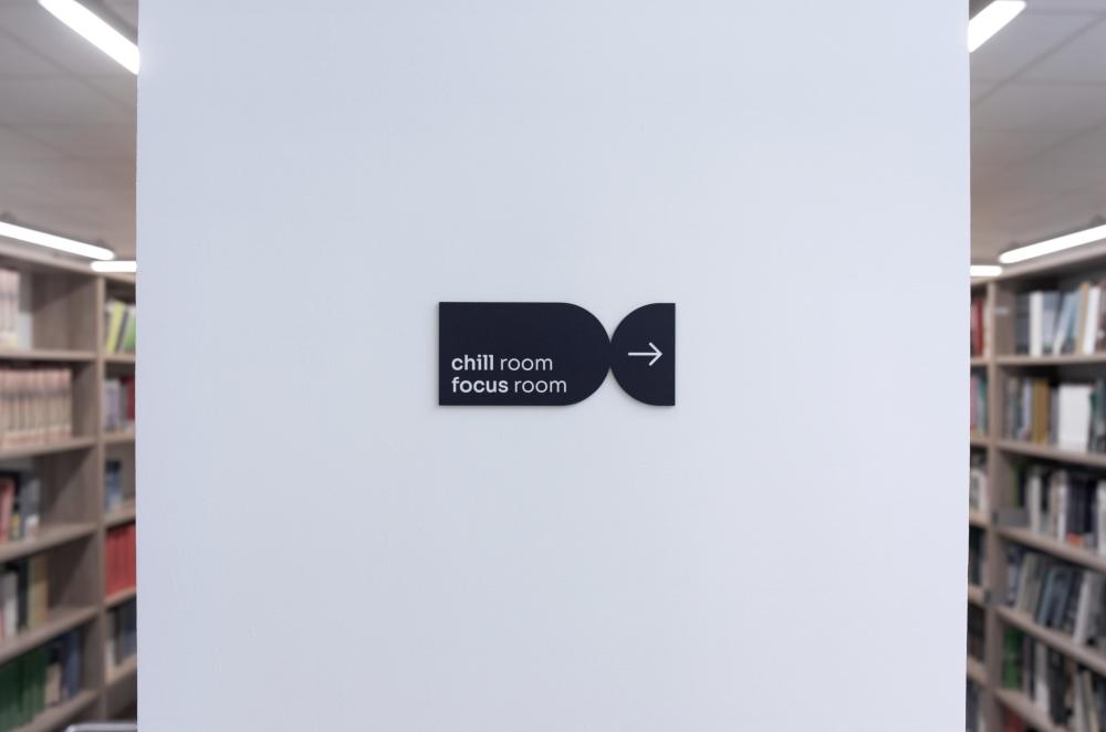 Black, horizontal directional sign on a white pillar with the text "chill room / focus room" and an arrow pointing right, with library bookshelves in the background.