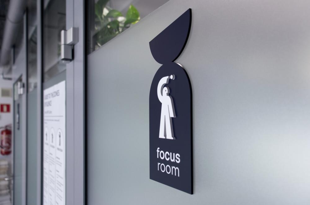Side view of a dark blue sign on glass featuring a white pictogram of a figure holding a dot over their head and the text "focus room".