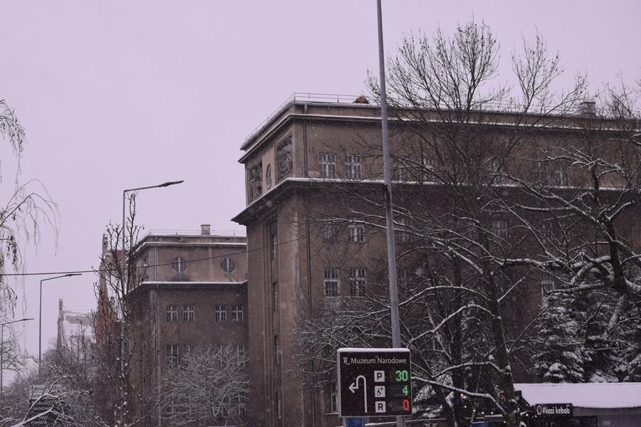 A perspective of the Silesian Seminary from a nearby vantage point. The scene encompasses two wings of the building, snow-laden trees, a glimpse of a bus stop, and a section of a road sign pointing the way to the National Museum.