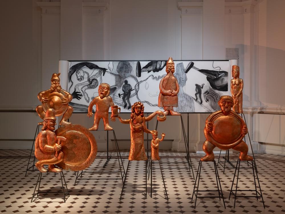 An art installation in a bright exhibition hall. Copper, exaggerated figures referencing the blacksmithing trade are displayed on thin black metal stands. In the background hangs a large-scale, black-and-white drawing featuring expressive sketches of people and animals at work.