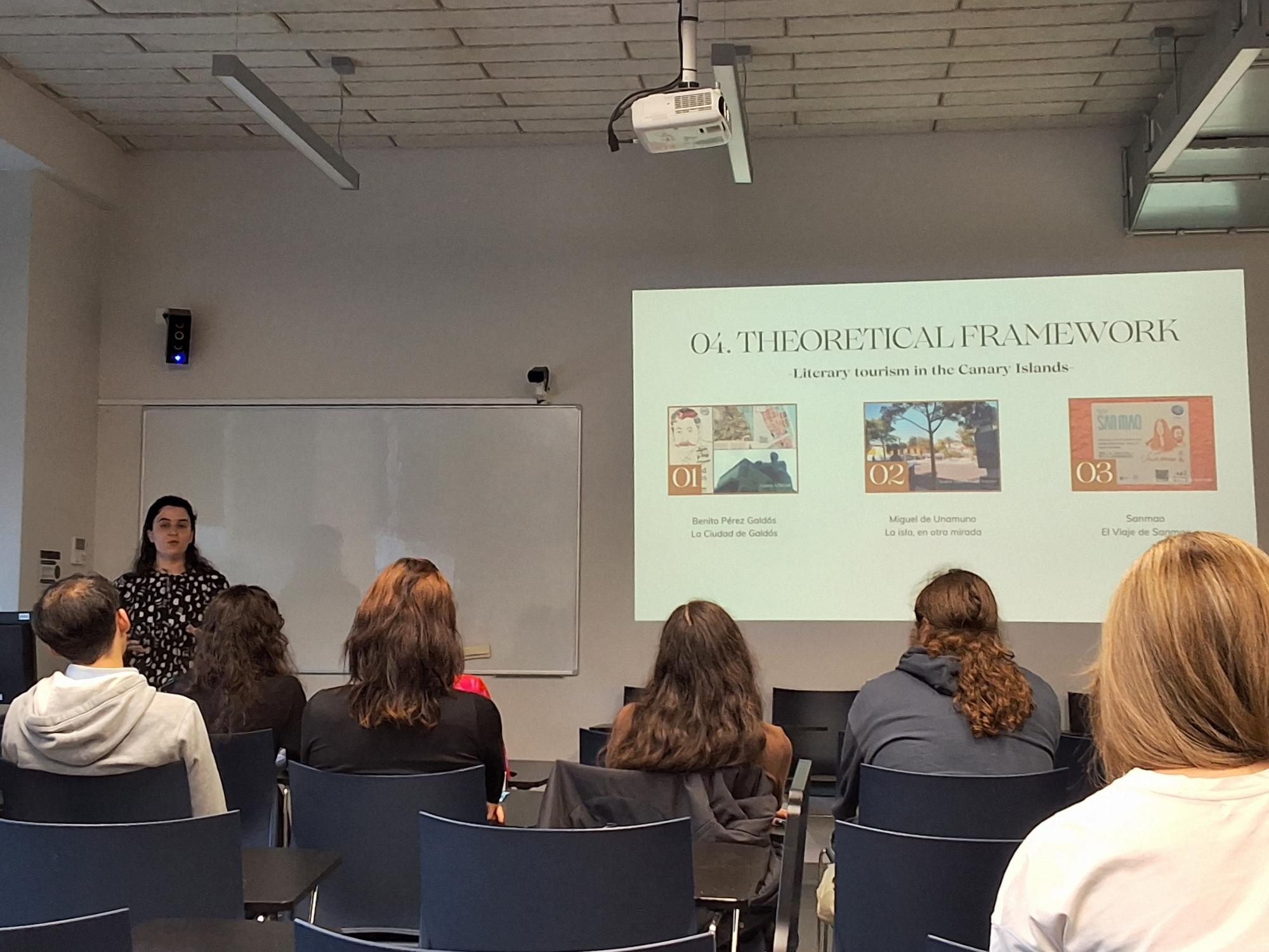 Conference audience seated in rows, viewed from behind, watching a speaker presenting at the front of the room beside a projected slide.