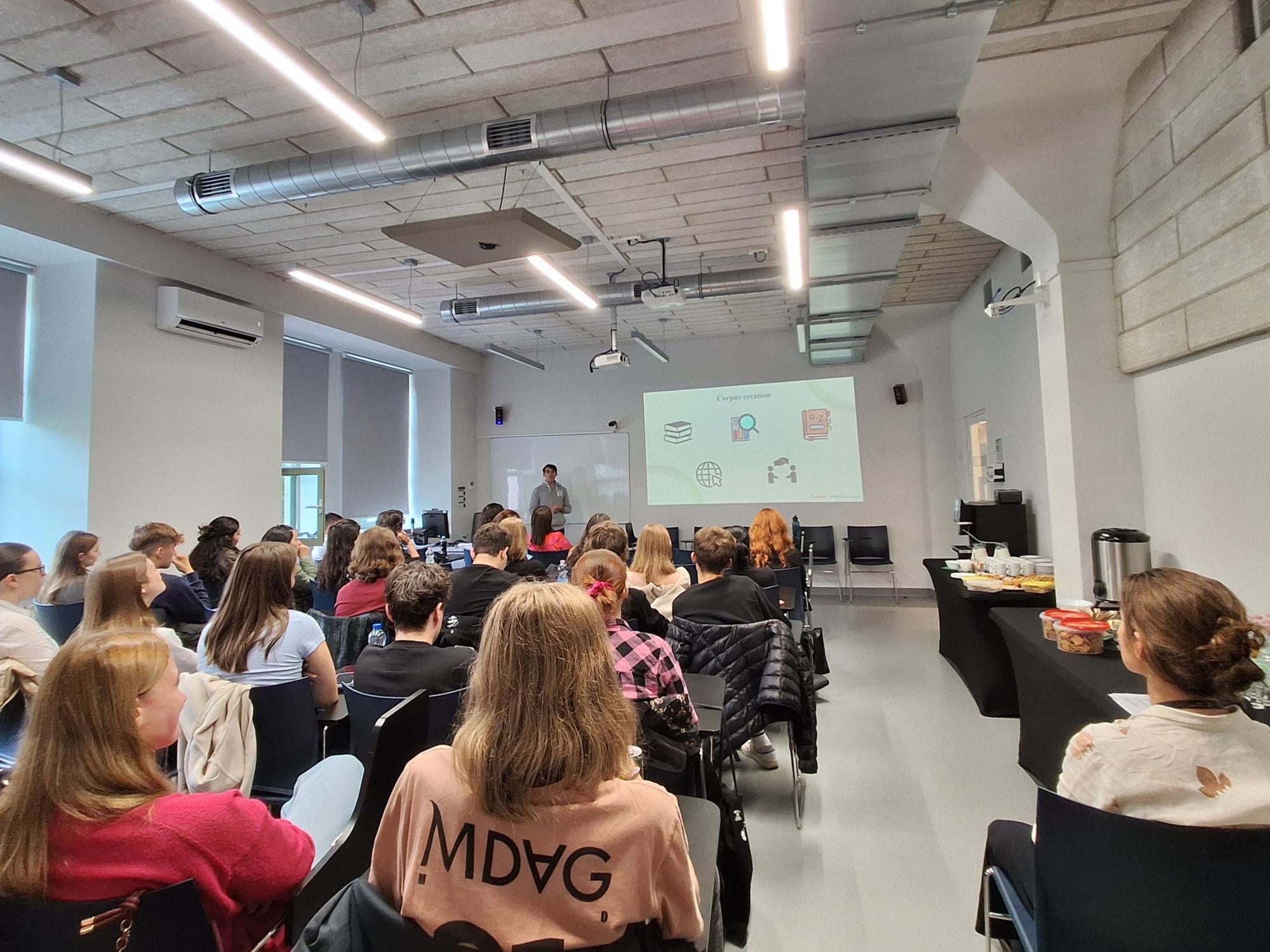 Conference audience seated in rows, viewed from behind, watching a speaker presenting at the front of the room beside a projected slide.