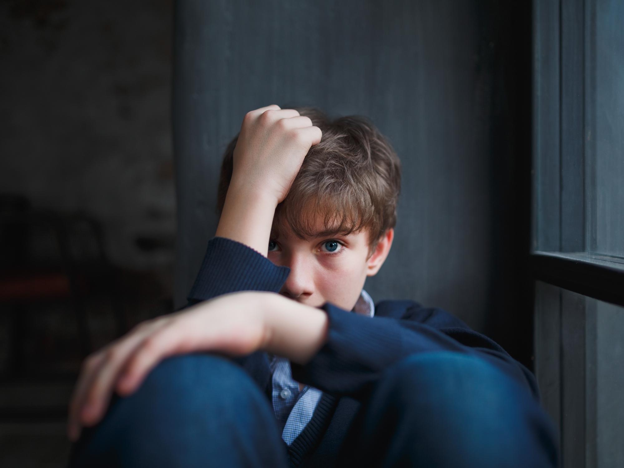 A troubled boy sitting by the window covers his face with his hand