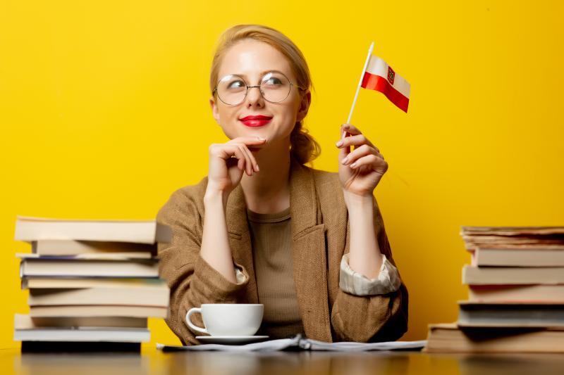 Smiling young woman in glasses sits at a desk flanked by piles of books